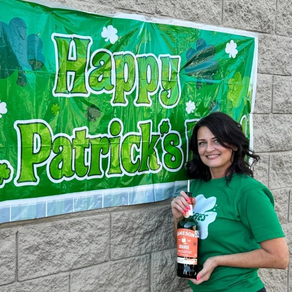 Woman in a green Wolfies shirt holding a Jameson Orange bottle in front of a "Happy Patrick's Day" banner.
