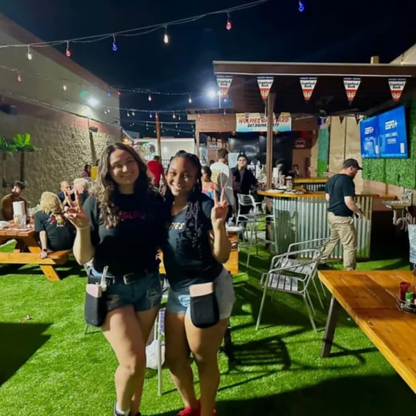 Two women pose with peace signs in an outdoor sports bar with string lights and patrons.