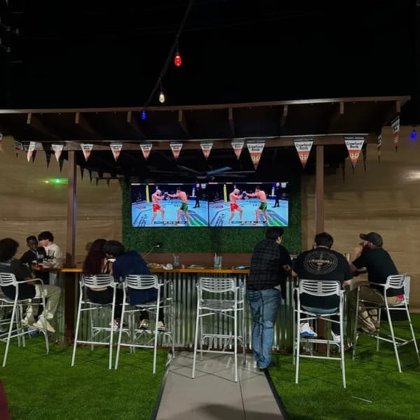 Patrons at Wolfies Sports Bar watch a fight on large screens in an outdoor seating area with artificial turf.