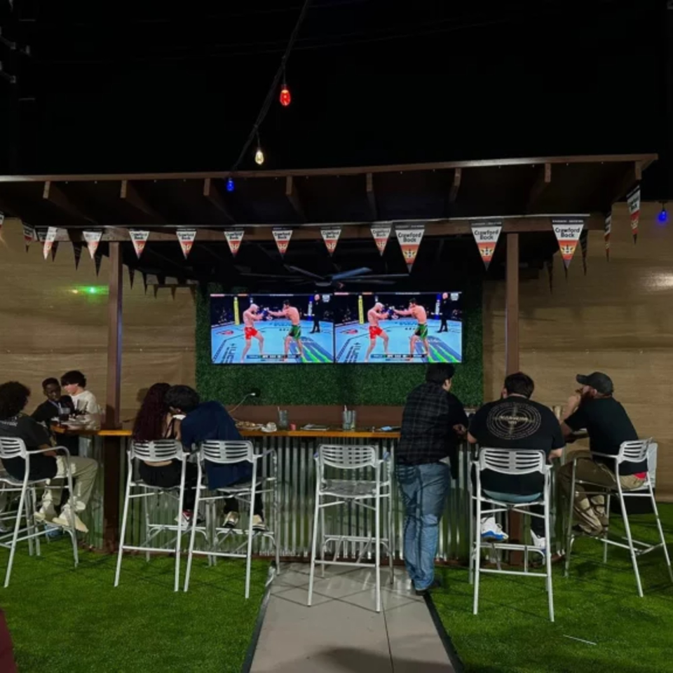Patrons at Wolfies Sports Bar watch a fight on large screens in an outdoor seating area with artificial turf.