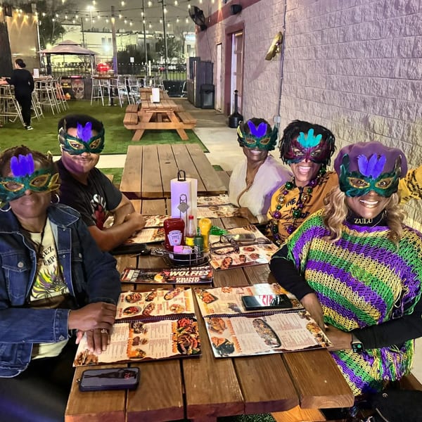 Five people in Mardi Gras masks and attire sit at a picnic table on Wolfies Restaurant patio.