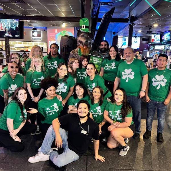 Wolfies staff in green St. Patrick's Day shirts pose with a wolf mascot in a sports bar.