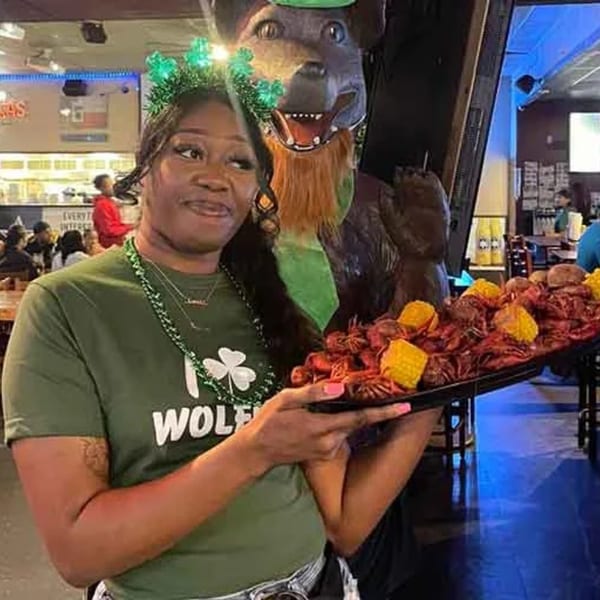 A woman in a Wolfies t-shirt holds a tray of crawfish and corn, next to a dog statue.