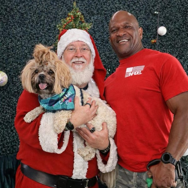 Santa Claus holding two small dogs, posing with a smiling man in a red shirt.