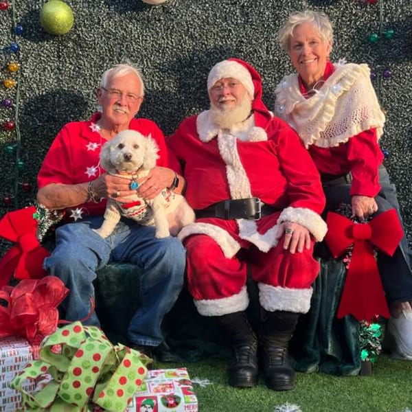 Santa Claus, a man with a dog, and a woman pose for a festive Christmas photo.