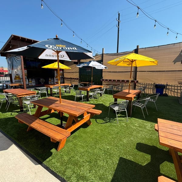 Outdoor patio with picnic tables, chairs, and umbrellas on artificial turf under a clear sky.
