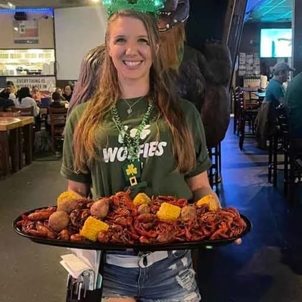 A smiling Wolfies server holds a large tray of crawfish, corn, and potatoes in a lively sports bar.