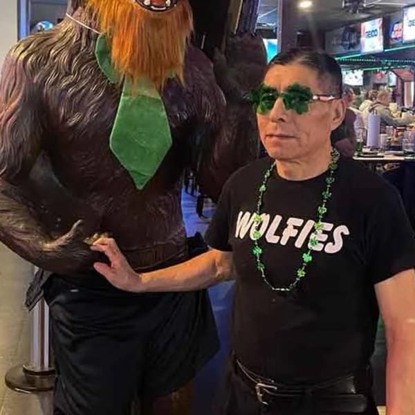 Man in Wolfies shirt and shamrock glasses poses with a Bigfoot statue wearing a green tie.