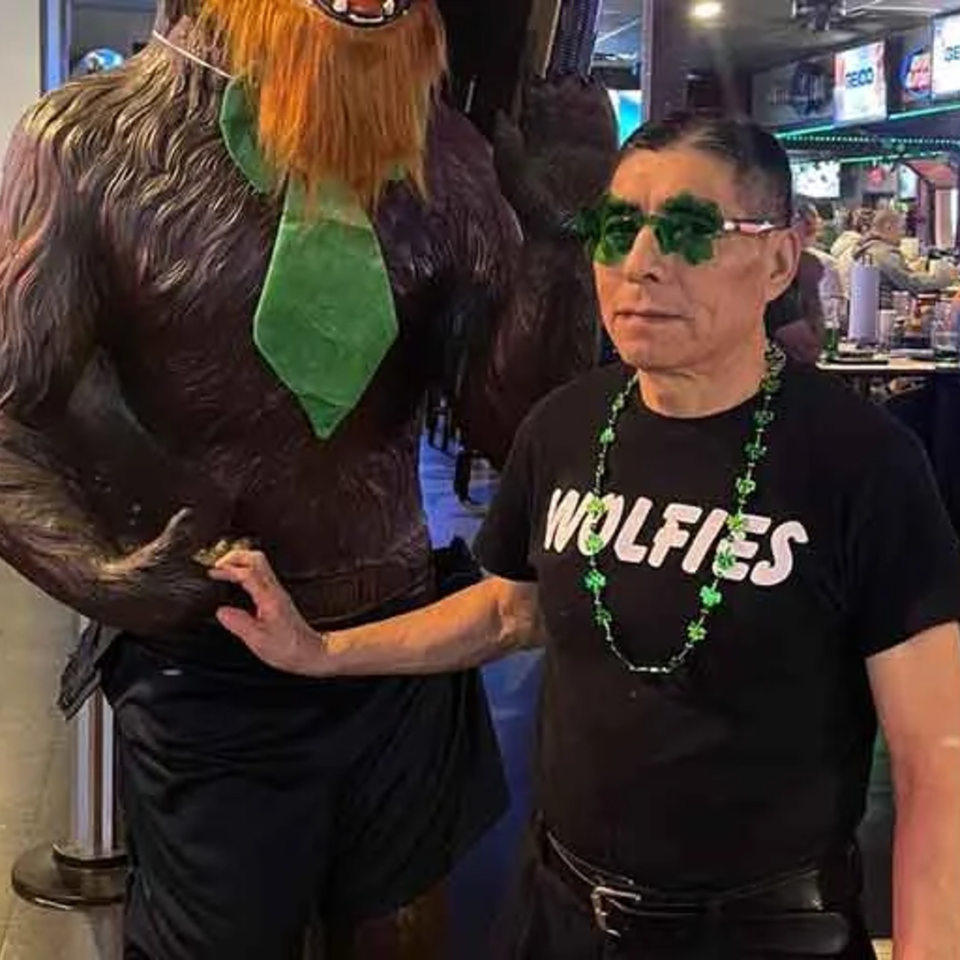 Man in Wolfies shirt and shamrock glasses poses with a Bigfoot statue wearing a green tie.