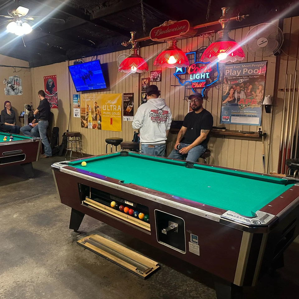 Patrons play pool at Wolfies Sports Bar, with neon signs and sports memorabilia adorning the walls.