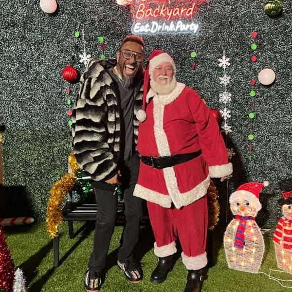 A man in a fur coat and Santa Claus pose smiling in front of a Christmas-decorated wall.