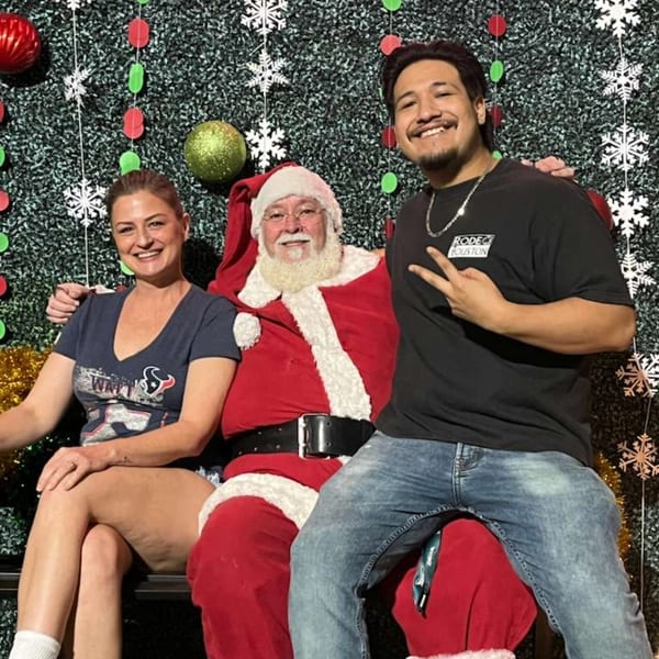 A woman and man pose with Santa Claus in front of a festive green backdrop with ornaments.