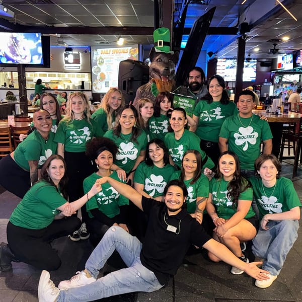 Wolfies staff in green shirts pose with a wolf mascot and "Happy St. Patrick's Day" sign.