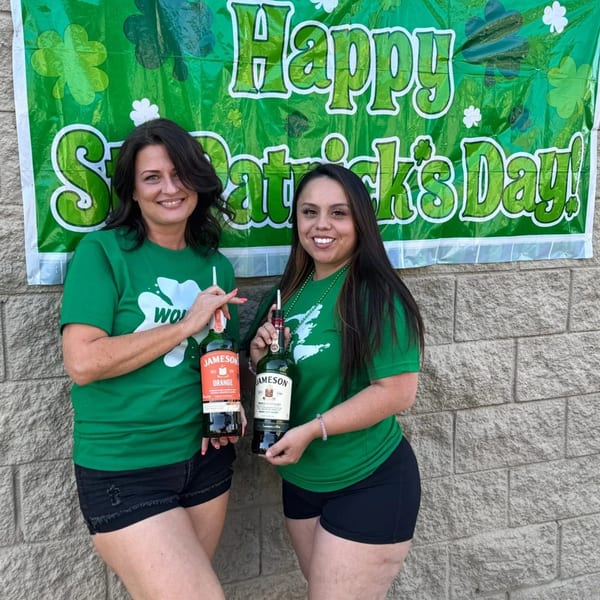 Two women in green shirts holding Jameson bottles in front of a "Happy St. Patrick's Day" banner.