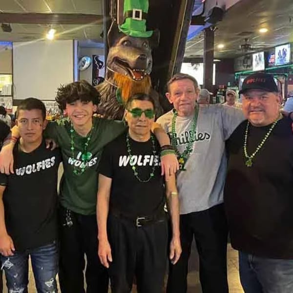 Five men in Wolfies shirts and green beads pose with a wolf statue wearing a leprechaun hat.