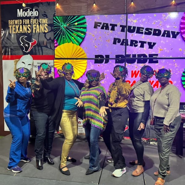 Seven women in Mardi Gras masks and festive attire pose in front of a "Fat Tuesday Party" sign.