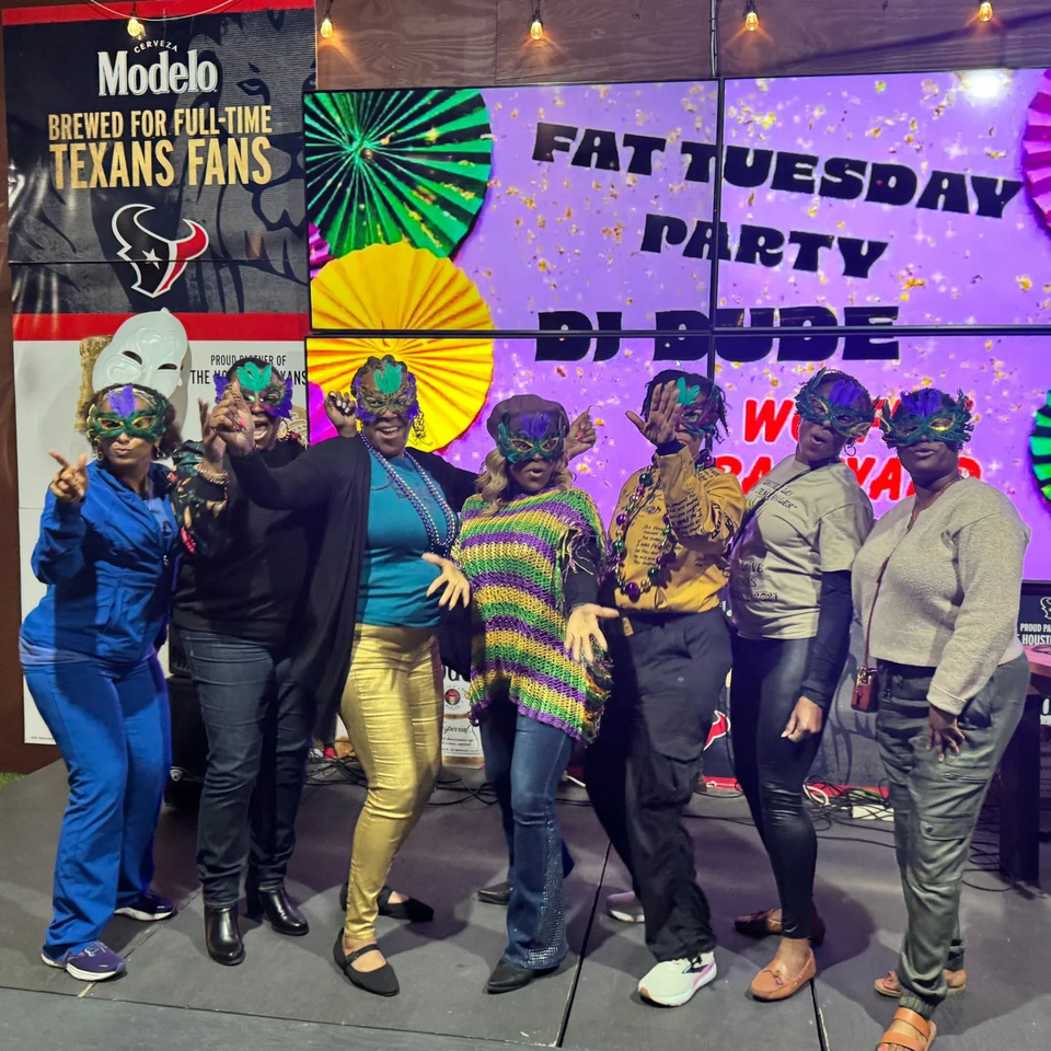 Seven women in Mardi Gras masks and festive attire pose in front of a "Fat Tuesday Party" sign.
