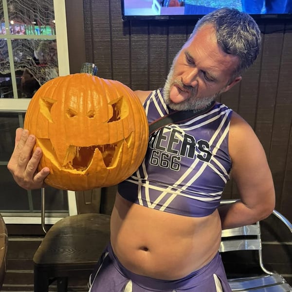 Man in a purple cheerleader outfit holding a carved Halloween pumpkin, sticking out his tongue.