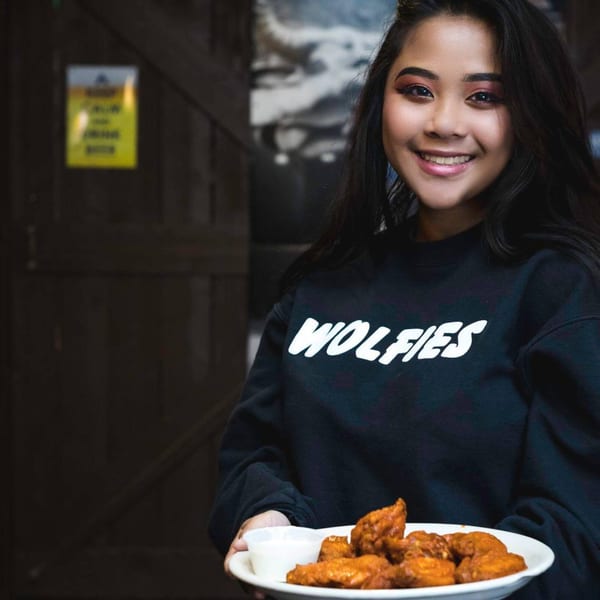 Smiling woman in a Wolfies sweatshirt holding a plate of chicken wings with dipping sauce.
