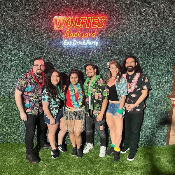 Six people in Hawaiian shirts and leis pose in front of a Wolfies Backyard neon sign.