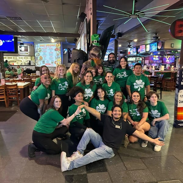 Wolfies staff in green shirts posing with a dog statue wearing a St. Patrick's Day hat.