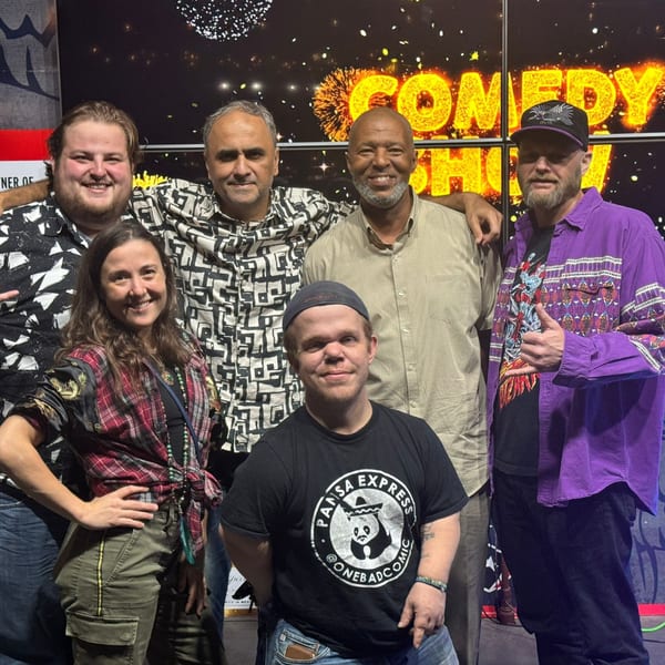 Six people smiling at the camera in front of a "Comedy Show" sign at Wolfies Sports Bar.