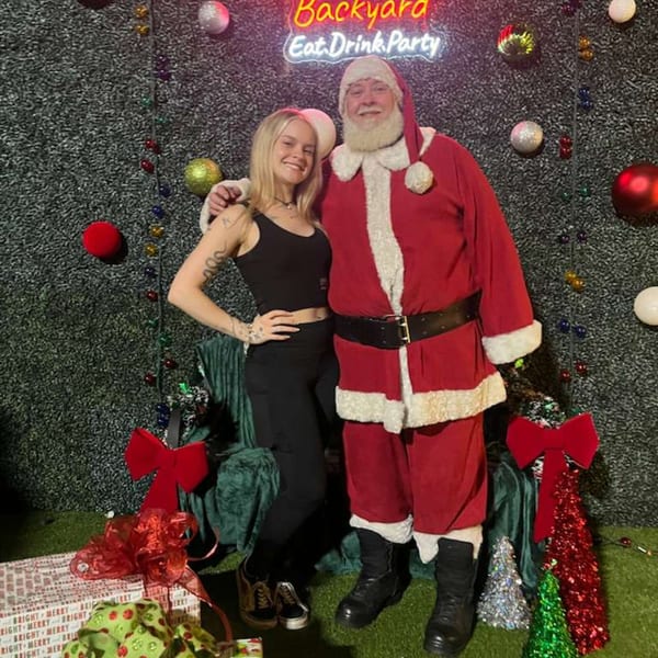 A woman and Santa Claus pose in front of a Christmas-themed backdrop with a neon sign.