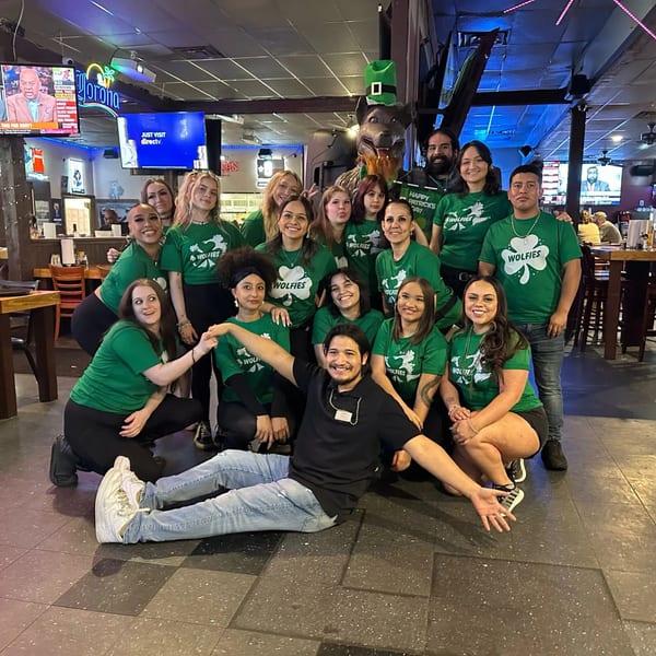 Wolfies Restaurant staff in green shirts with shamrocks, celebrating St. Patrick's Day in a sports bar.