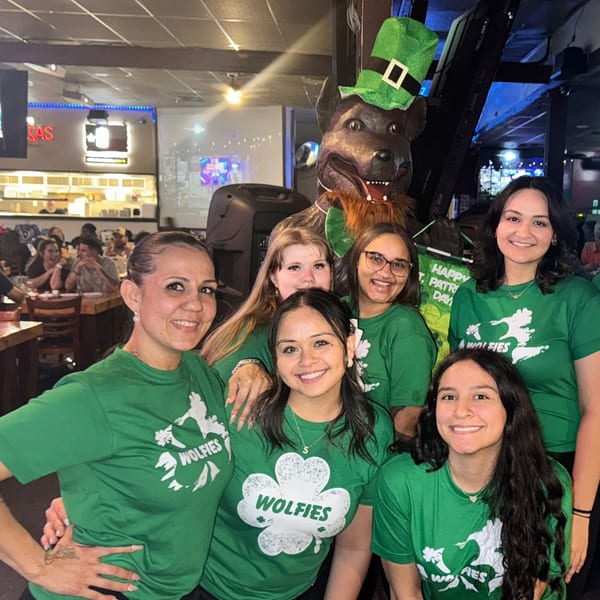 Six smiling Wolfies staff in green shirts with a dog statue wearing a leprechaun hat.
