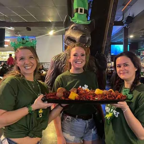Three smiling women in green shirts holding a large tray of crawfish, corn, and potatoes at Wolfies.