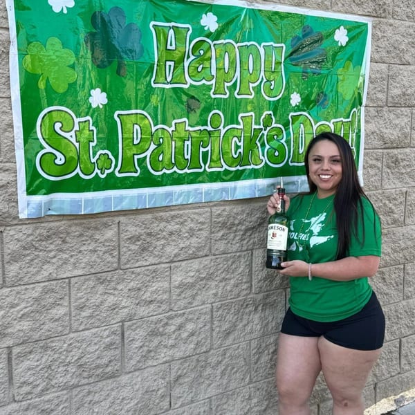 Woman in green Wolfies shirt holding a Jameson bottle in front of a "Happy St. Patrick's Day" banner.
