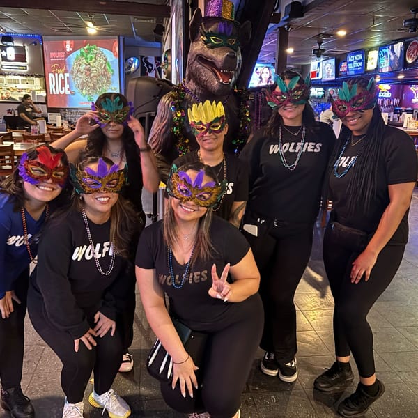 Wolfies staff in Mardi Gras masks pose with a large wolf mascot in the sports bar.