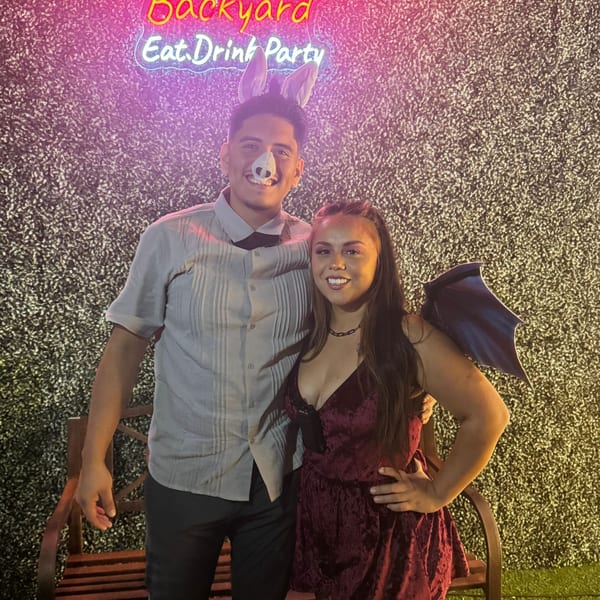 A man in a pig nose and bunny ears with a woman in devil wings pose in front of a neon sign.