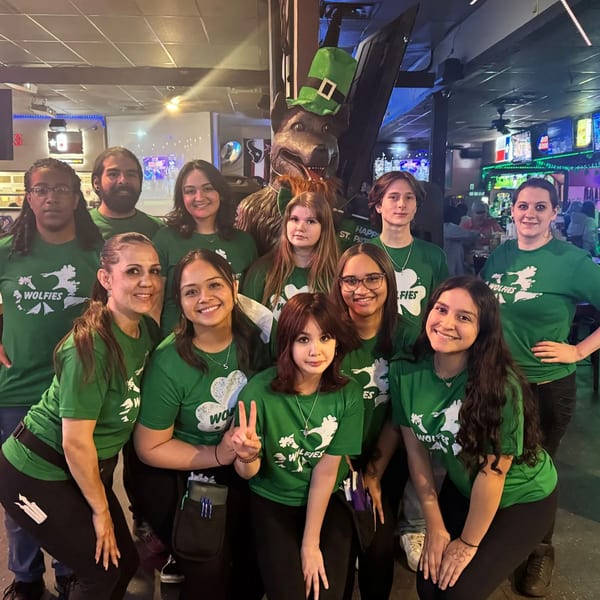 Wolfies staff in green shirts pose with a wolf statue wearing a leprechaun hat for St. Patrick's Day.