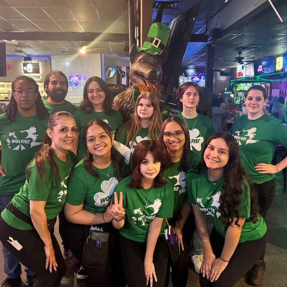 Wolfies staff in green shirts pose with a wolf statue wearing a leprechaun hat for St. Patrick's Day.
