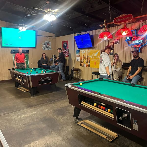 Patrons playing pool and socializing in a dimly lit sports bar with neon signs and TVs.