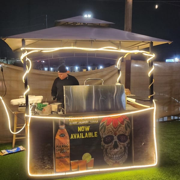 A vendor stands inside a brightly lit outdoor bar stall with a "Mango Shotta Now Available" sign.