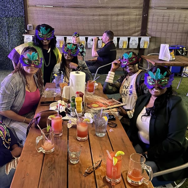 Five women in colorful Mardi Gras masks and a man drinking at a wooden table in a sports bar.
