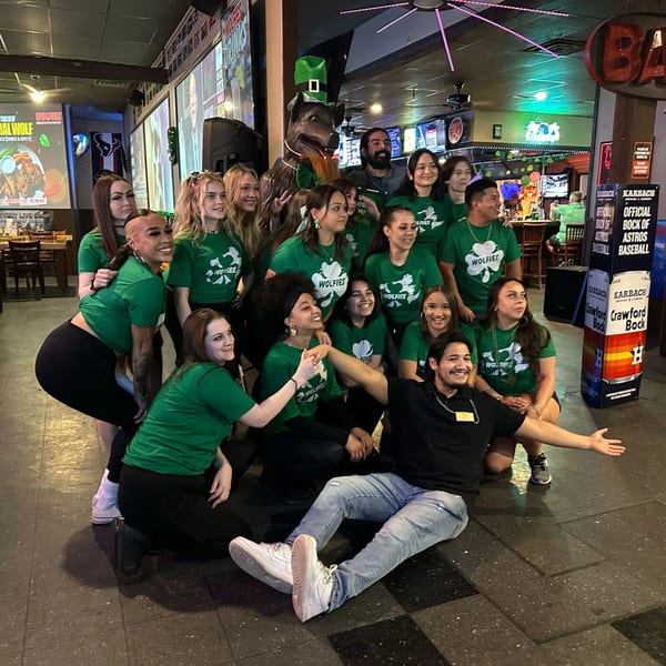 Wolfies Restaurant and Sports Bar staff in green shirts posing with a wolf statue wearing a leprechaun hat.