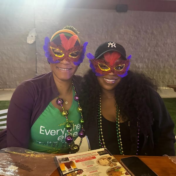 Two women in colorful Mardi Gras masks and beads smile at Wolfies Restaurant and Sports Bar.