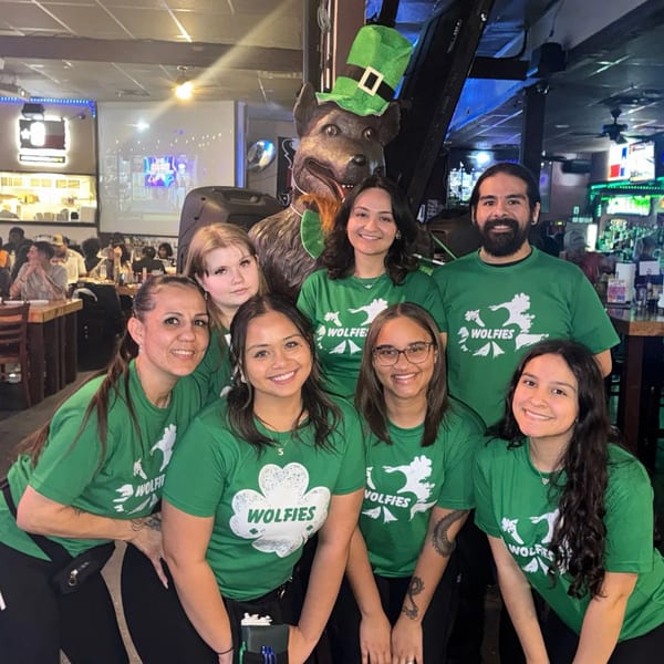 Wolfies staff in green shirts pose with a wolf statue wearing a St. Patrick's Day hat.
