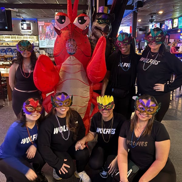 Seven women in Wolfies shirts and Mardi Gras masks pose with a giant inflatable lobster and wolf.