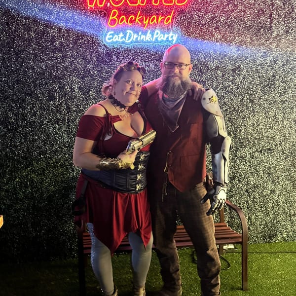 Two people in steampunk attire pose under a neon sign that reads "Wolfies Backyard Eat. Drink. Party."