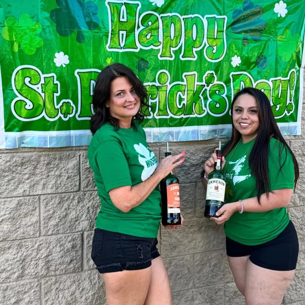 Two women in green shirts holding Jameson bottles in front of a "Happy St. Patrick's Day" banner.