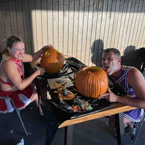 Two people in sports-themed costumes carving pumpkins at a table, with one person sticking out their tongue.