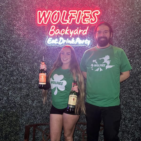 Two Wolfies staff members in green shirts holding Jameson bottles under a neon sign.