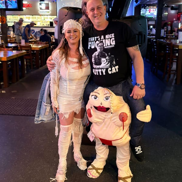 A couple in Halloween costumes, a mummy and a man riding a doll, smiling in a sports bar.