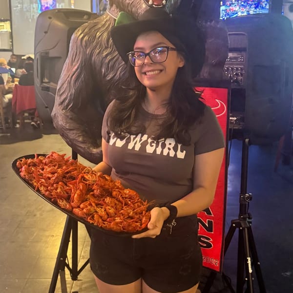 A smiling woman in a cowboy hat and "Cowgirl" shirt holds a large tray of crawfish.