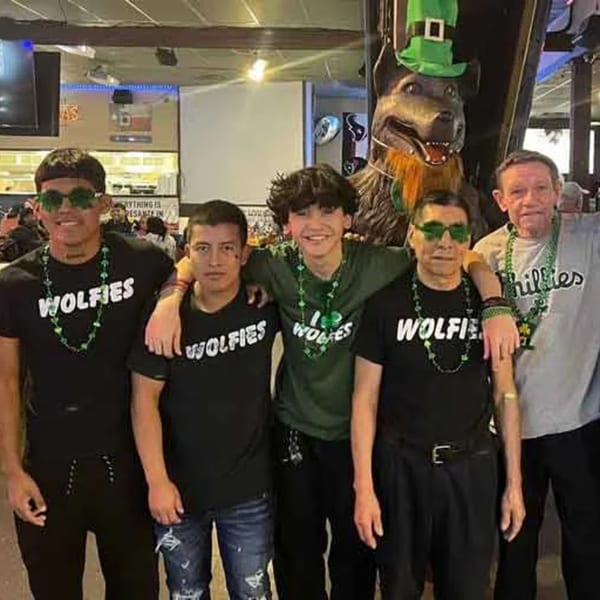 Five men wearing Wolfies shirts and green St. Patrick's Day beads pose in a sports bar.