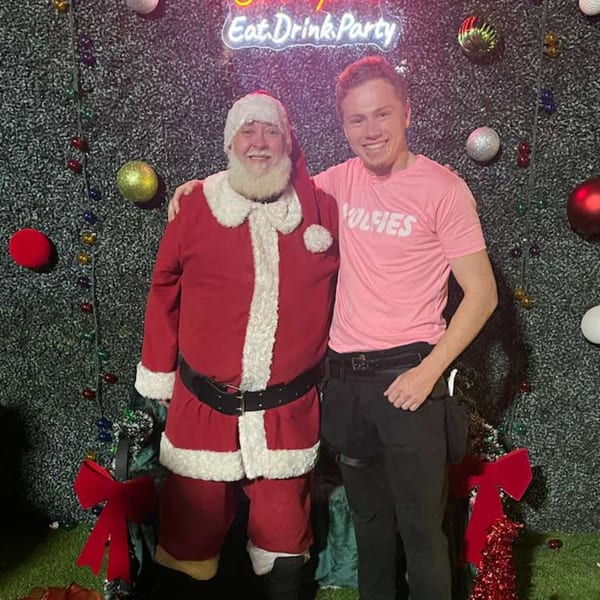 Santa Claus and a Wolfies employee in a pink shirt pose in front of a festive holiday wall.